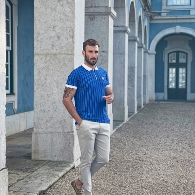 Homem com t-shirt azul riscada e calças claras em pátio com pilares de pedra e paredes azuis