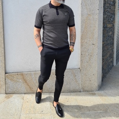 Homem com camisa polo riscada cinzenta e preta, calças pretas e sapatos pretos na calçada