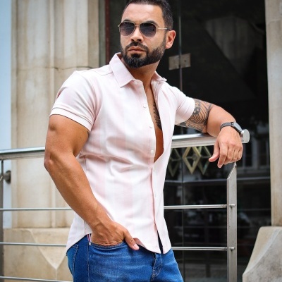 Homem com camisa rosa clara e calças de ganga em ambiente urbano.