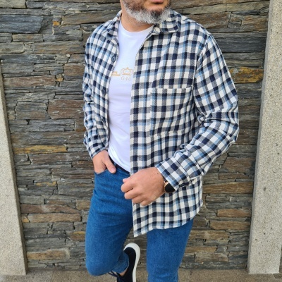 Homem com camisa xadrez azul e branca e jeans junto a parede de pedra