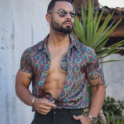 Homem com camisa colorida de padrão geométrico, calças pretas e óculos escuros junto a plantas e parede branca.
