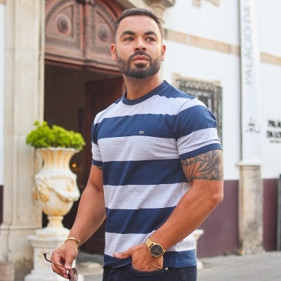 Homem de t-shirt às riscas azuis e cinza e jeans, com relógio dourado e óculos de sol na mão.