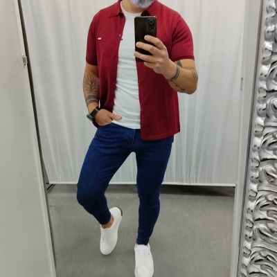 Homem com camisa vermelha, camisola branca, calças azuis e sapatilhas brancas a tirar selfie num espelho