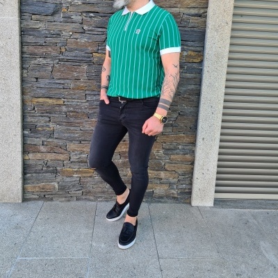 Homem com polo verde riscada e calças pretas em frente a parede de pedra