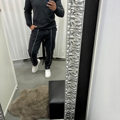 Homem a vestir conjunto desportivo preto com linhas brancas nas calças e sapatilhas brancas refletido em espelho com moldura prateada.