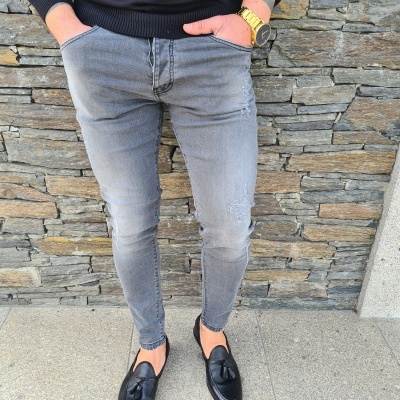 Pessoa vestindo calças de ganga cinza, suéter preto, sapatos pretos com borlas e relógio dourado, em frente a parede de pedra cinzenta.