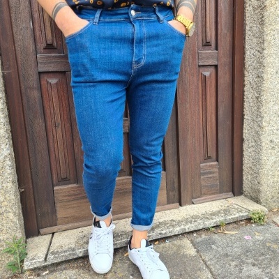 Calças de ganga azuis, camisa preta com padrão amarelo e tênis brancos em pavimento pedra diante de porta de madeira.