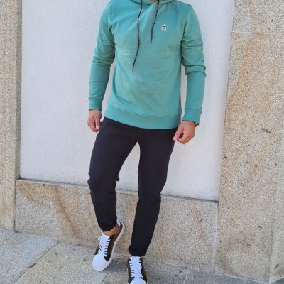 Homem com hoodie verde-água, calças pretas e ténis brancos perto de parede de pedra.