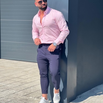 Homem em roupa casual com camisa rosa, calças azuis e sapatilhas brancas encostado numa parede cinza