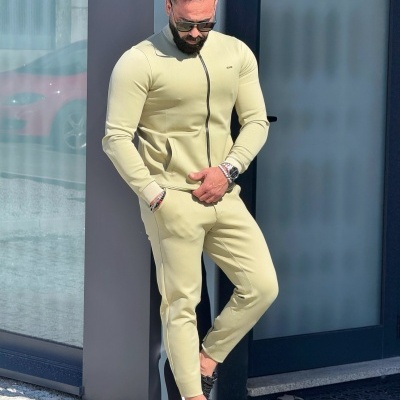 Homem com fato de treino bege claro encostado numa parede junto a porta de vidro