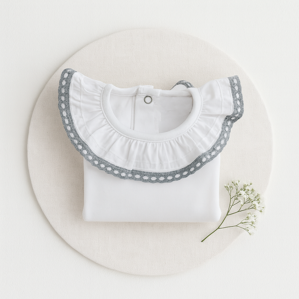 Camisola branca para bebé com gola de folho e bordado cinzento sobre base redonda branca com ramo de flores.