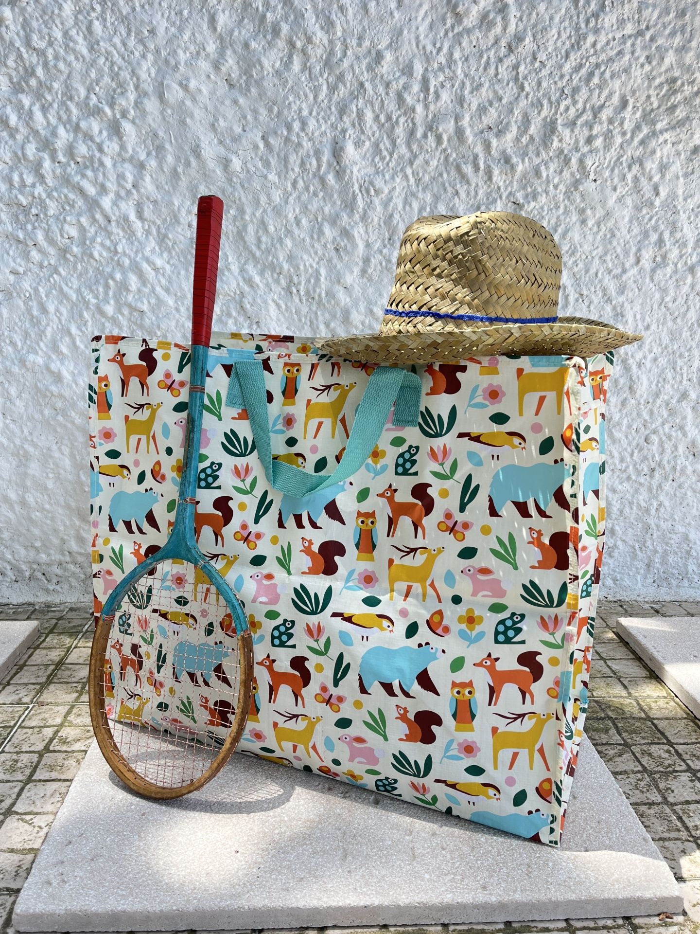 Saco com padrão de animais, chapéu de palha e raquete de madeira num chão de pedra
