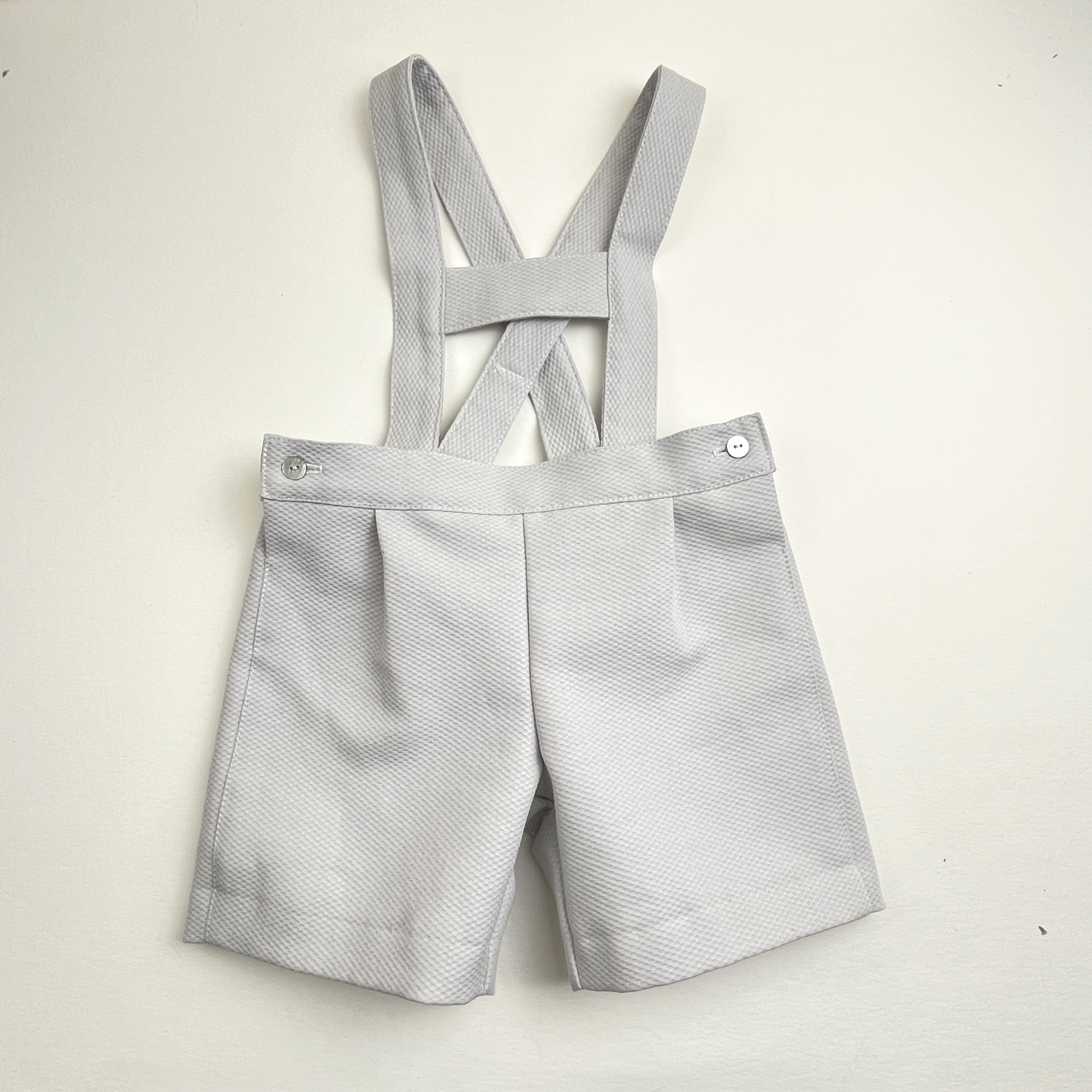 Calções cinzentos claros para criança com tirantes cruzados e botões prateados.