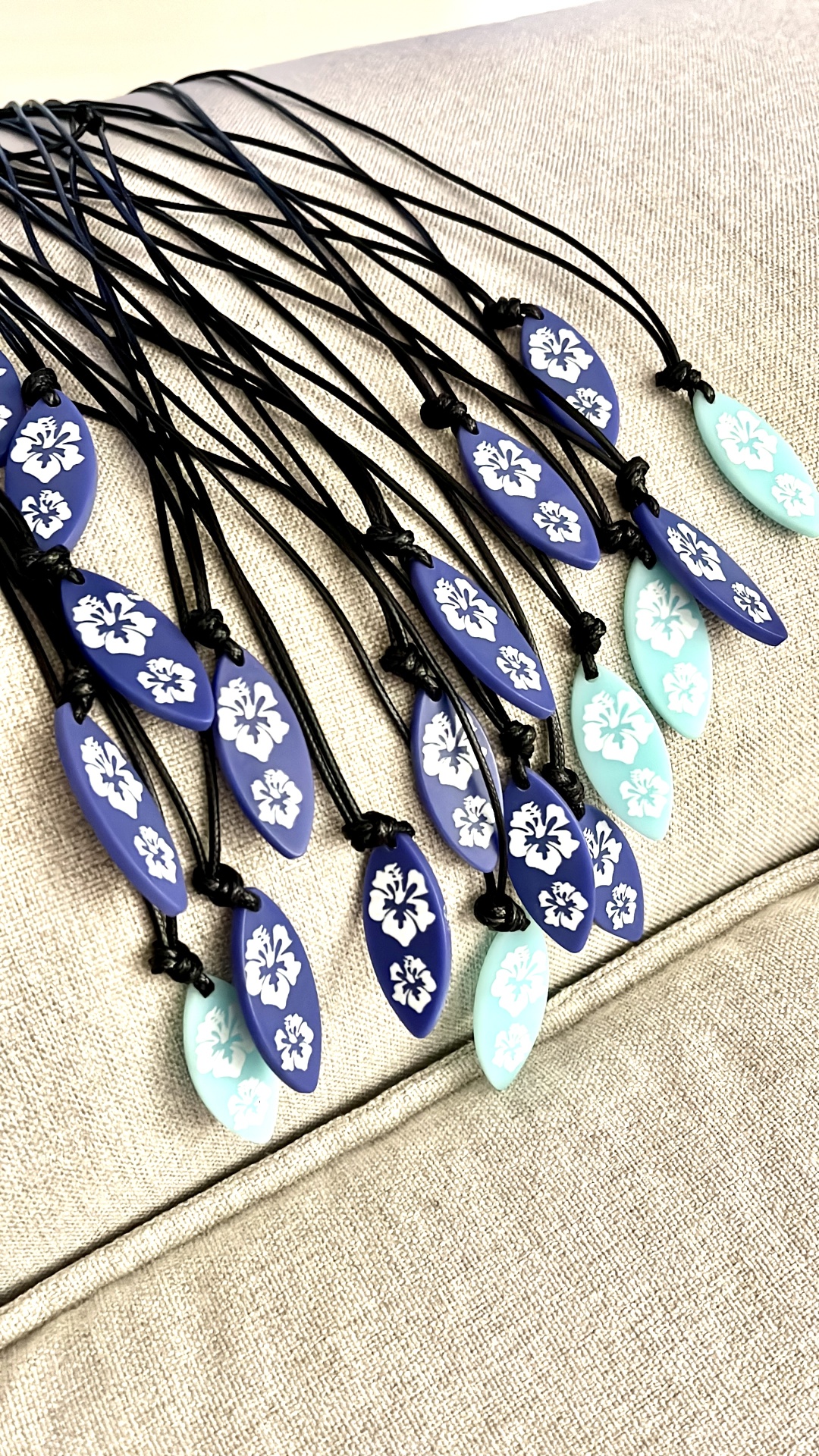 Colares com pendentes em formato de prancha azul e verde água com flores brancas sobre tecido cinzento claro