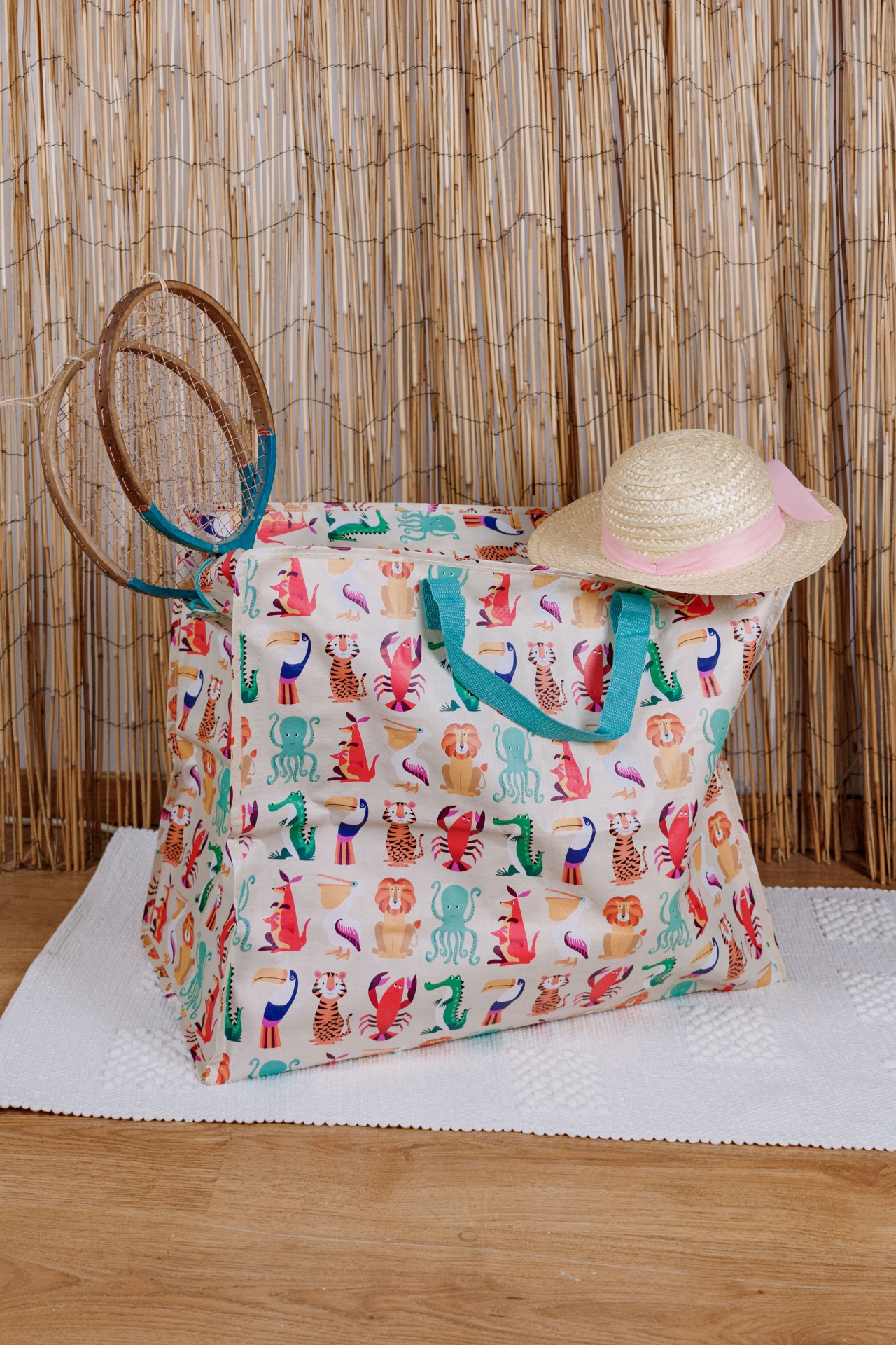 Saco com padrão de animais, chapéu de palha e raquetes ao lado, em ambiente com tapete branco e fundo de cana seca
