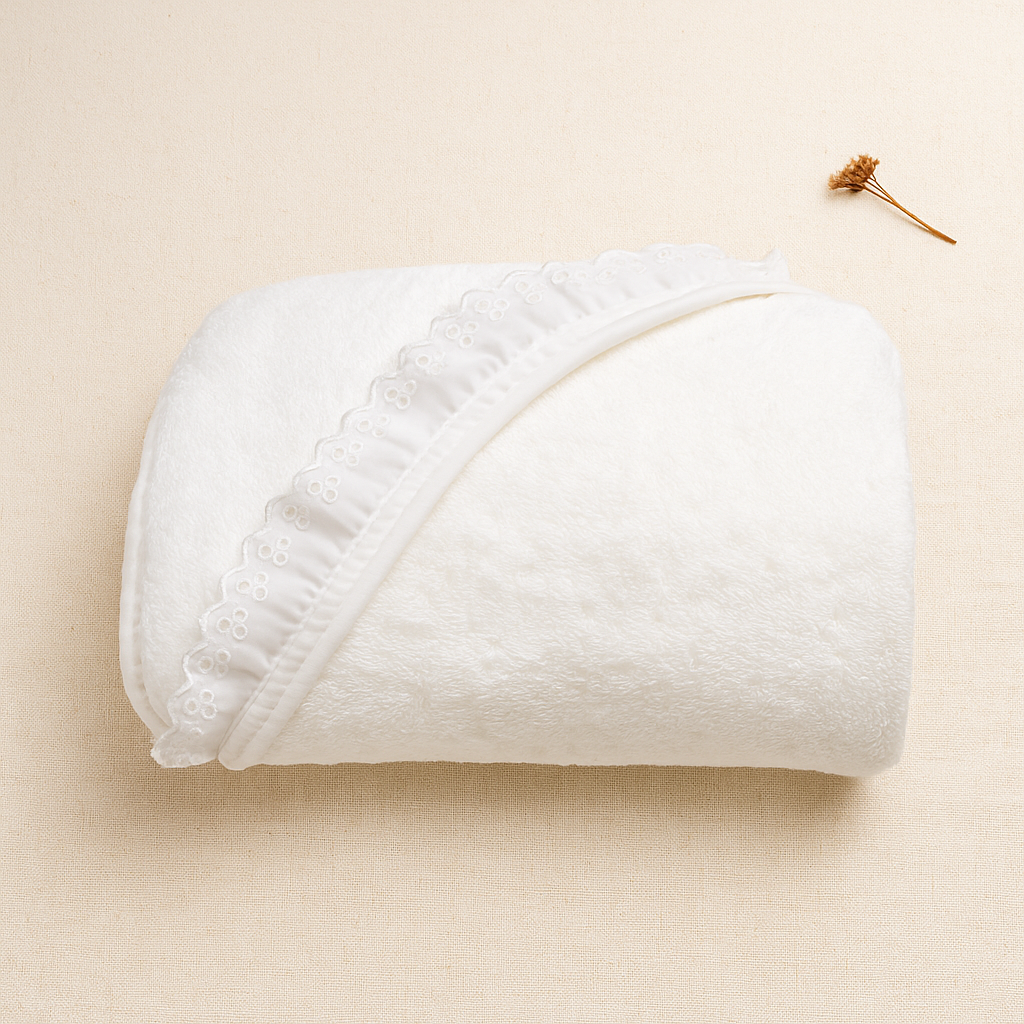 Toalha branca dobrada com capuz e bordado floral ao redor, sobre superfície bege com flor seca ao lado.