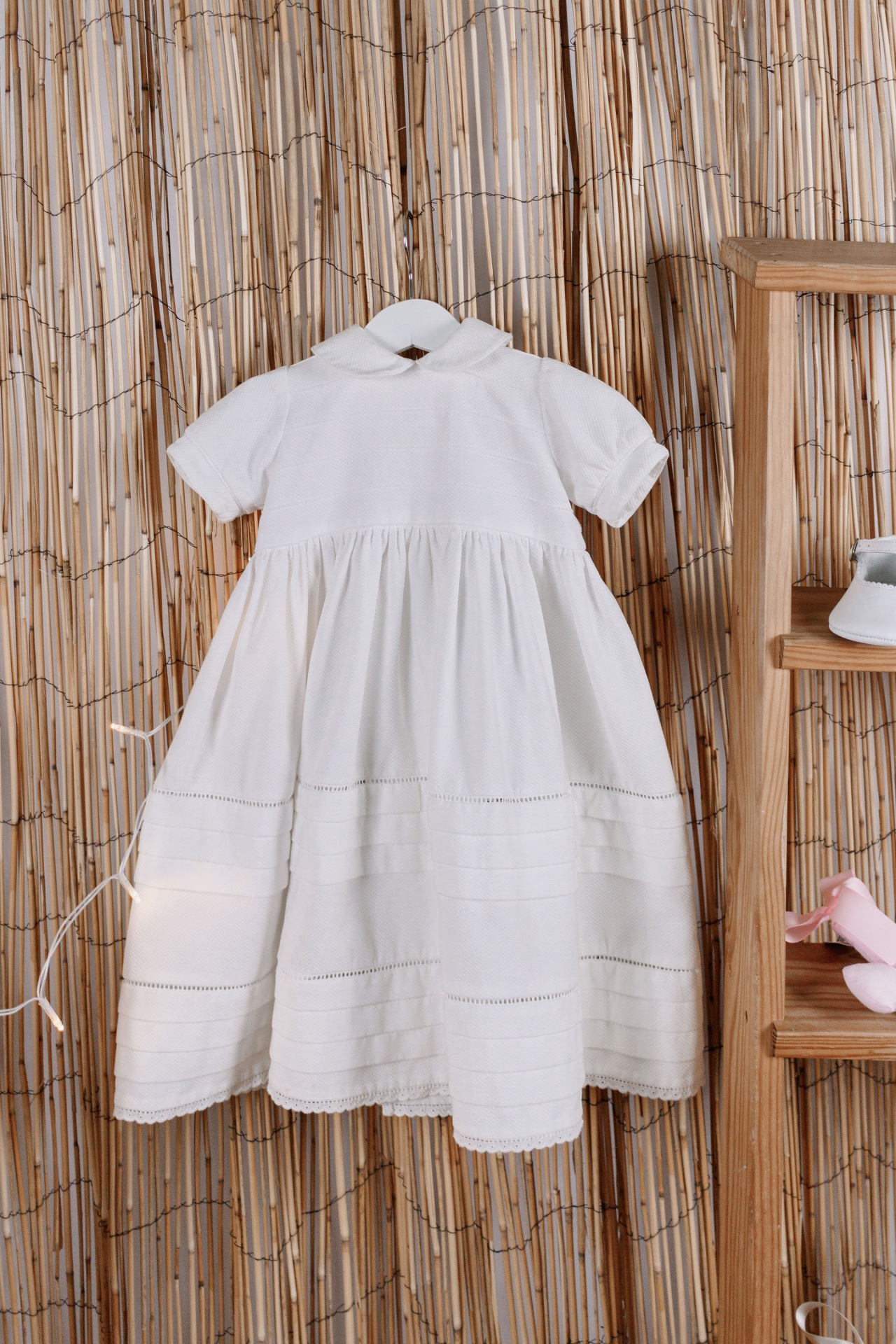 Vestido branco de bebé com gola peter pan pendurado contra fundo de palhinha