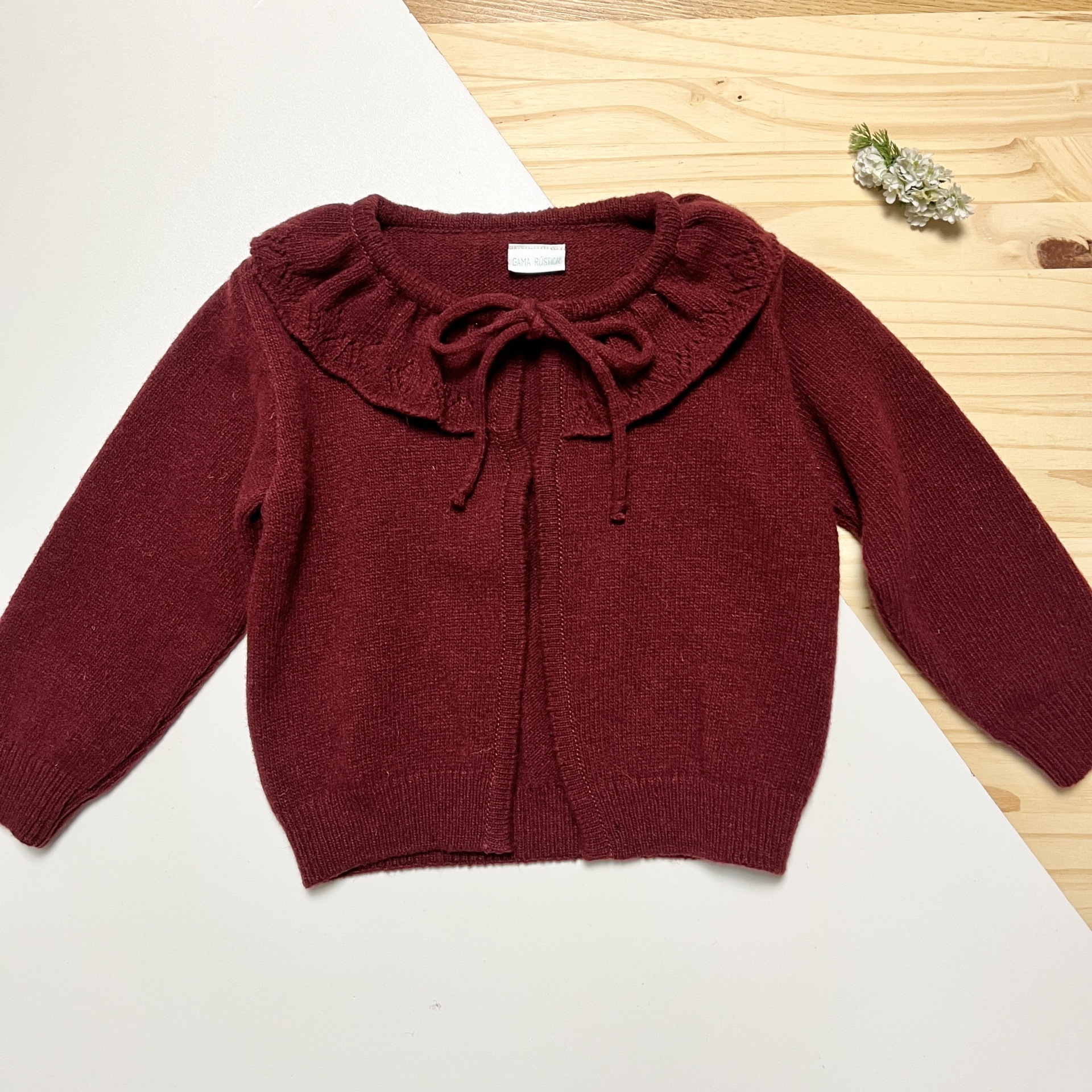 Camisola de malha vinho com gola folhada e laço frontal sobre fundo branco e madeira clara