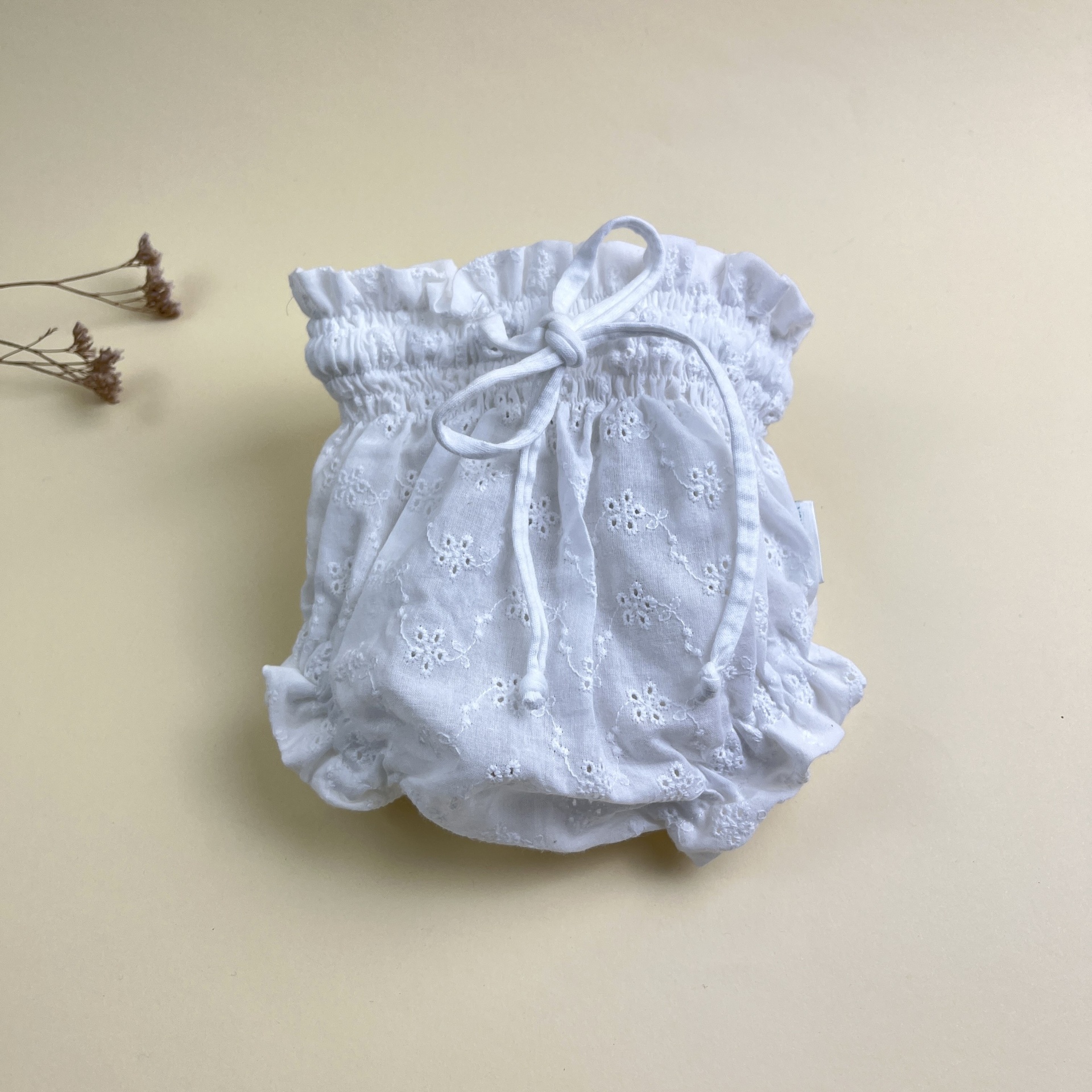 Culote branco para bebé com padrão broderie anglaise e fita ajustável, sobre fundo bege claro