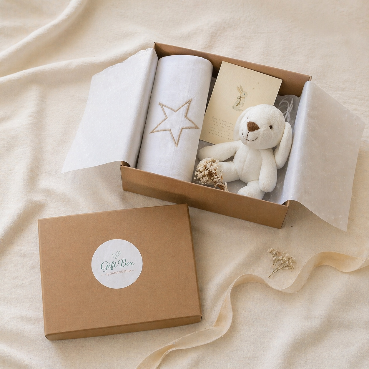 Caixa de presente com manta branca, peluche de coelho, cartão ilustrado e flores secas sobre tecido bege.