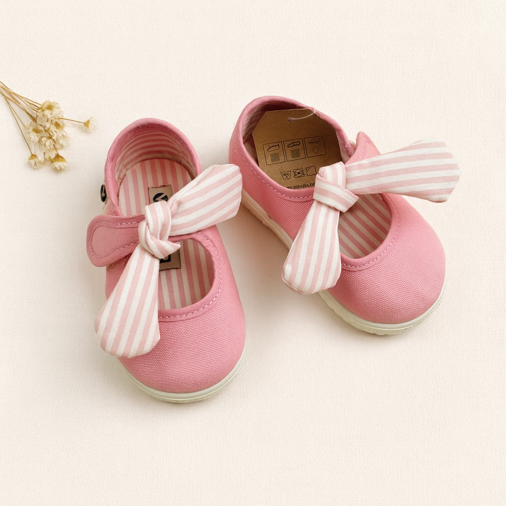 Sapatilhas cor-de-rosa para bebé com laços listrados e etiqueta no interior, sobre fundo claro com flores secas