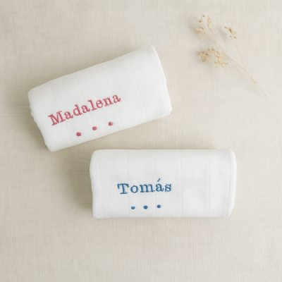 Dois toalhetes brancos com nomes bordados Madalena e Tomás, sobre fundo bege com ramo seco