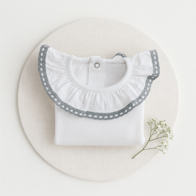 Camisola branca para bebé com gola de folho e bordado cinzento sobre base redonda branca com ramo de flores.