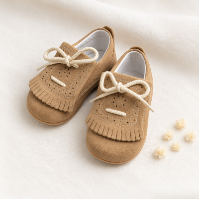 Sapatos em camurça para bebé marrons com franjas e cordões brancos sobre tecido branco.