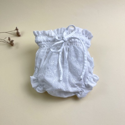 Culote branco para bebé com padrão broderie anglaise e fita ajustável, sobre fundo bege claro