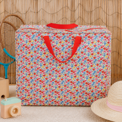Bolsa grande colorida com padrão floral e alças vermelhas ao lado de raquete, câmara de madeira e chapéu de palha.