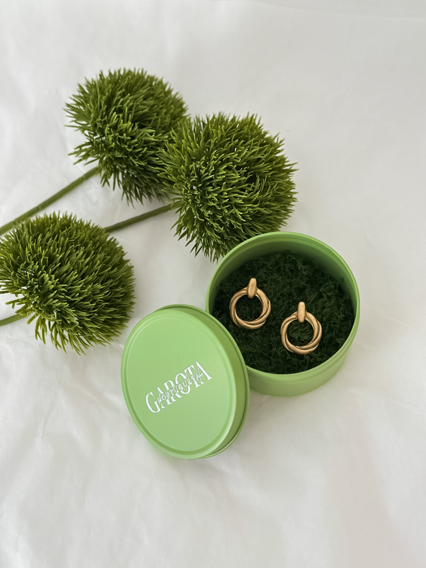 Aros Brincos dourados em caixa verde com raminhos decorativos verdes ao lado.