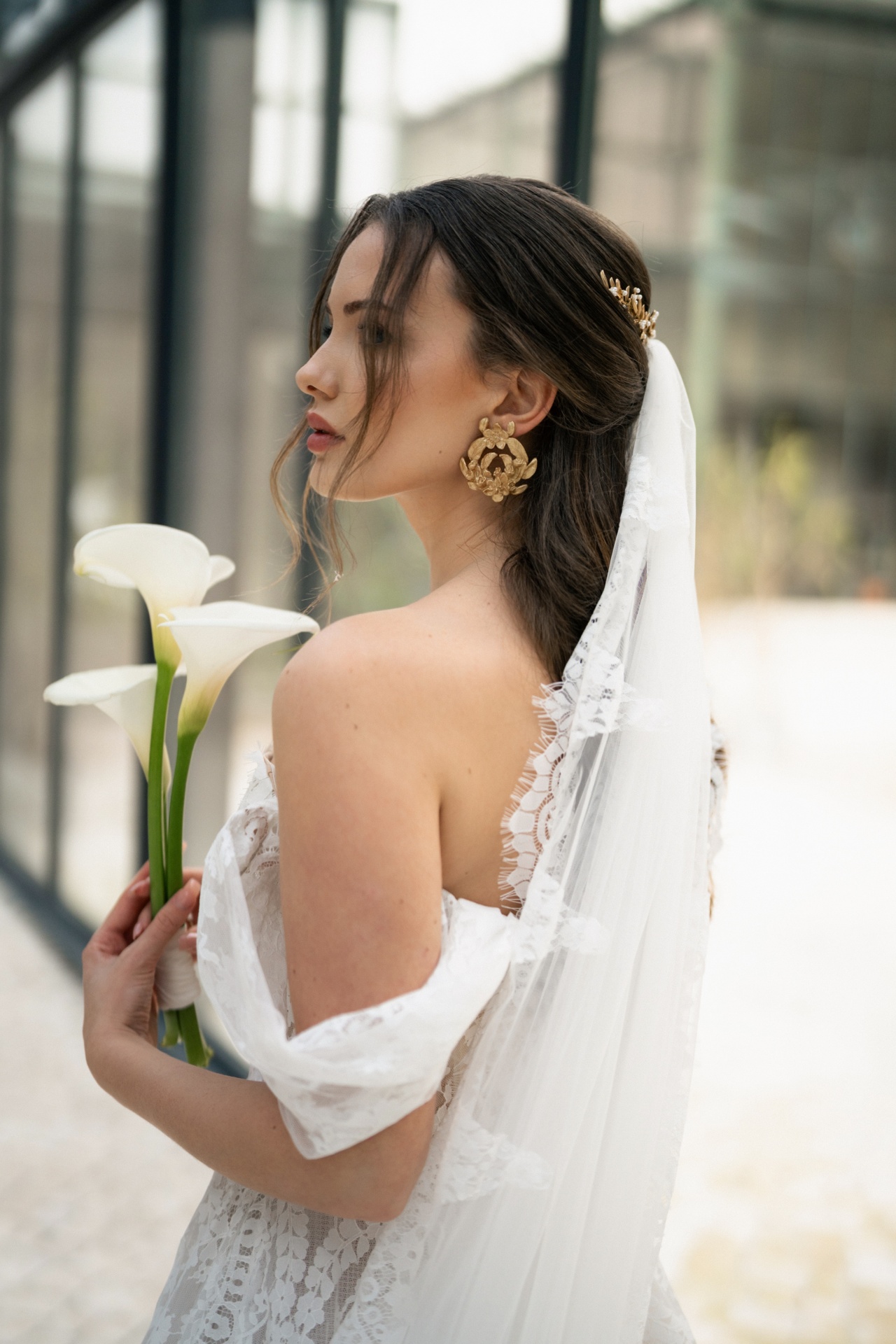 Erbária Noiva de perfil com vestido rendado branco, véu com renda, brincos dourados grandes e segurando flores brancas