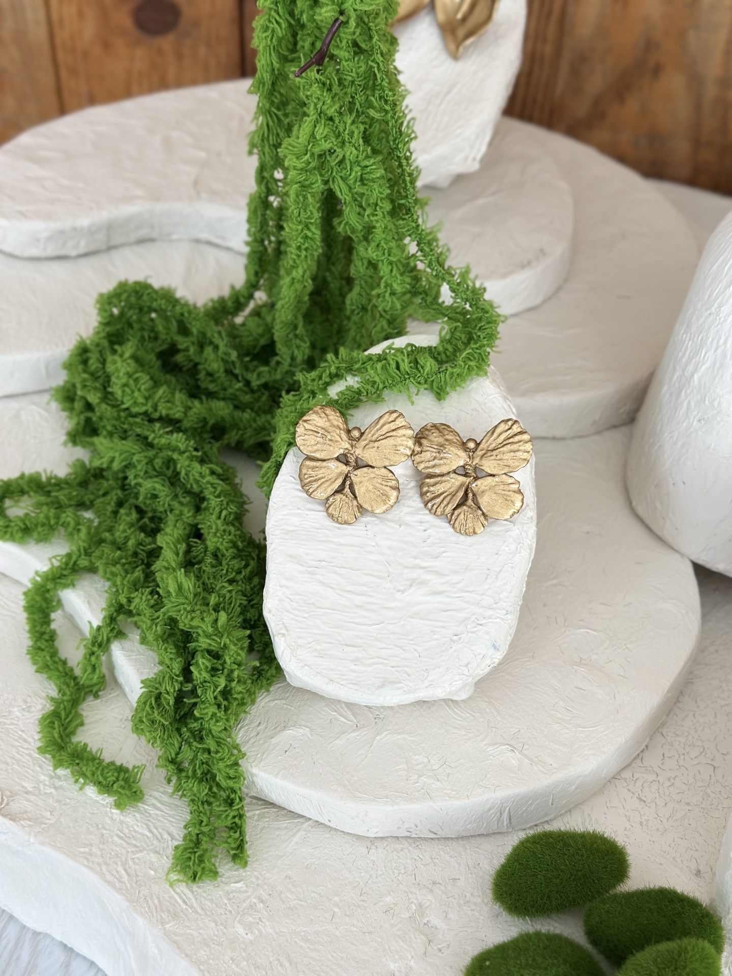 Foglia Brincos de flores douradas sobre pedestal branco com decoração verde ao redor.