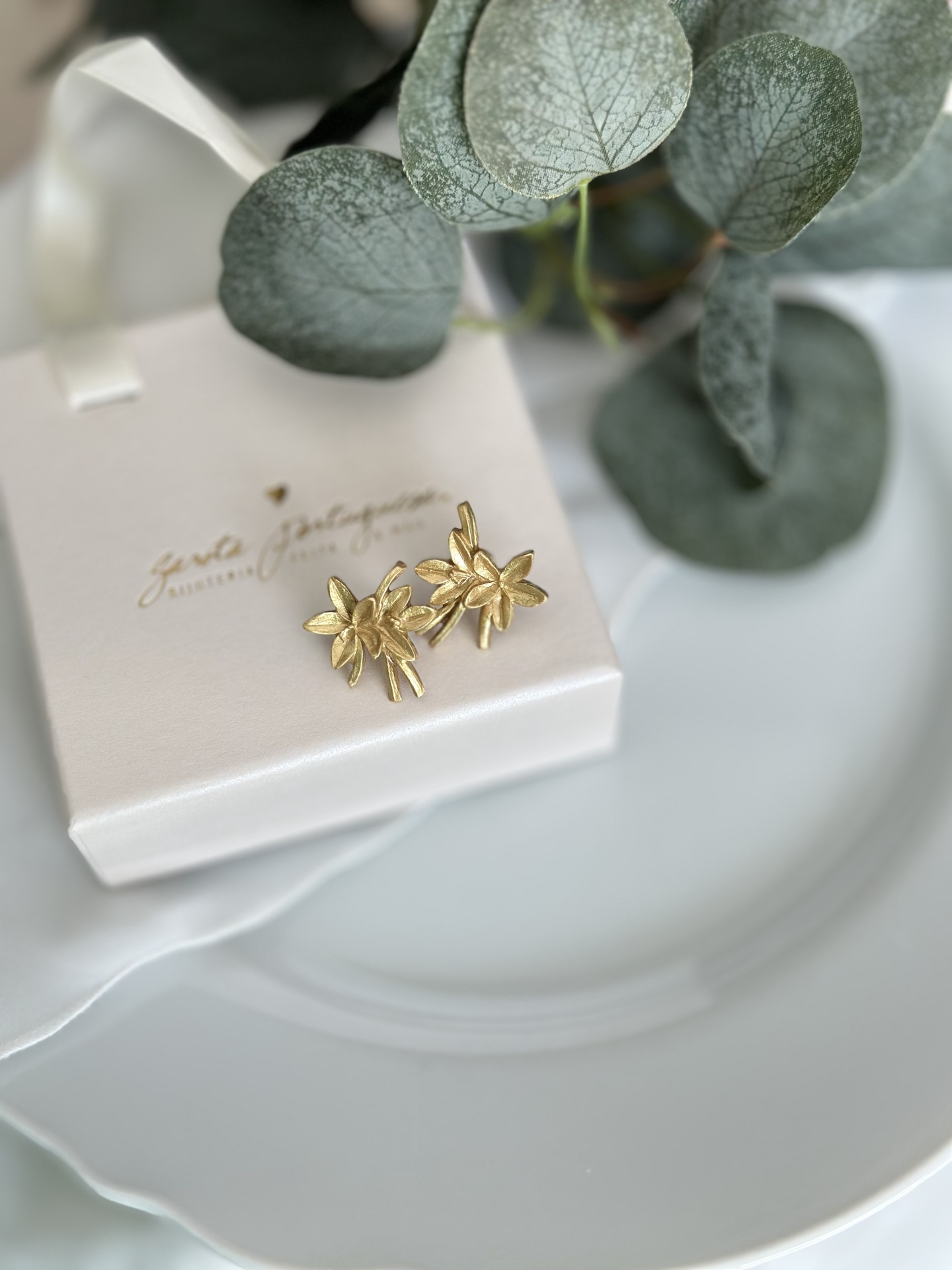 Zínia Brincos dourados em forma de flores sobre caixa branca com texto dourado e folhas verdes secas