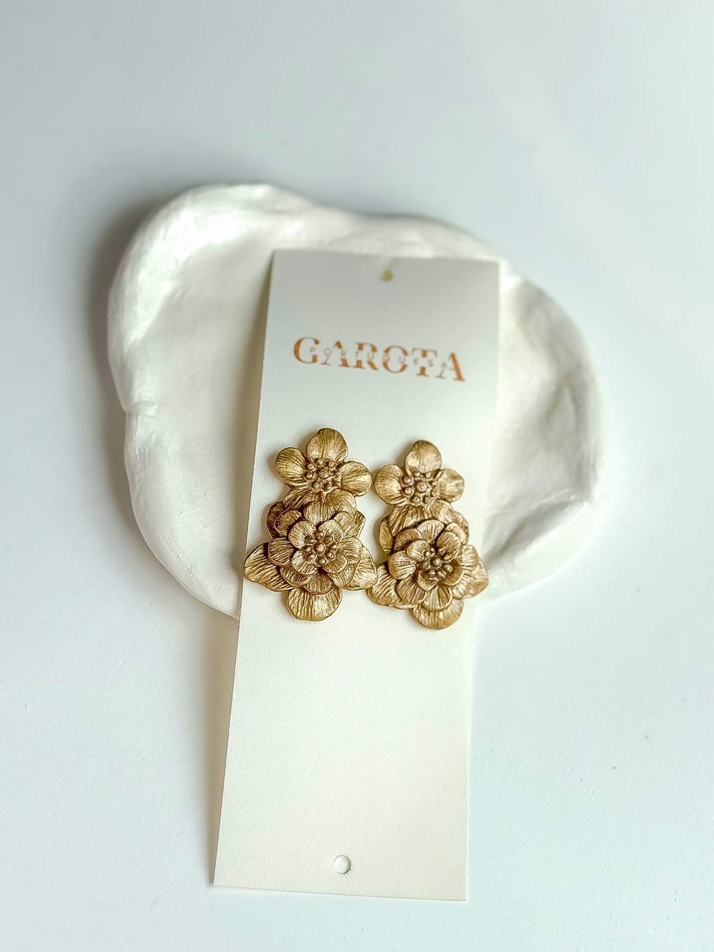 Madalena II brincos dourados em forma de flores num suporte branco com texto GAROTA