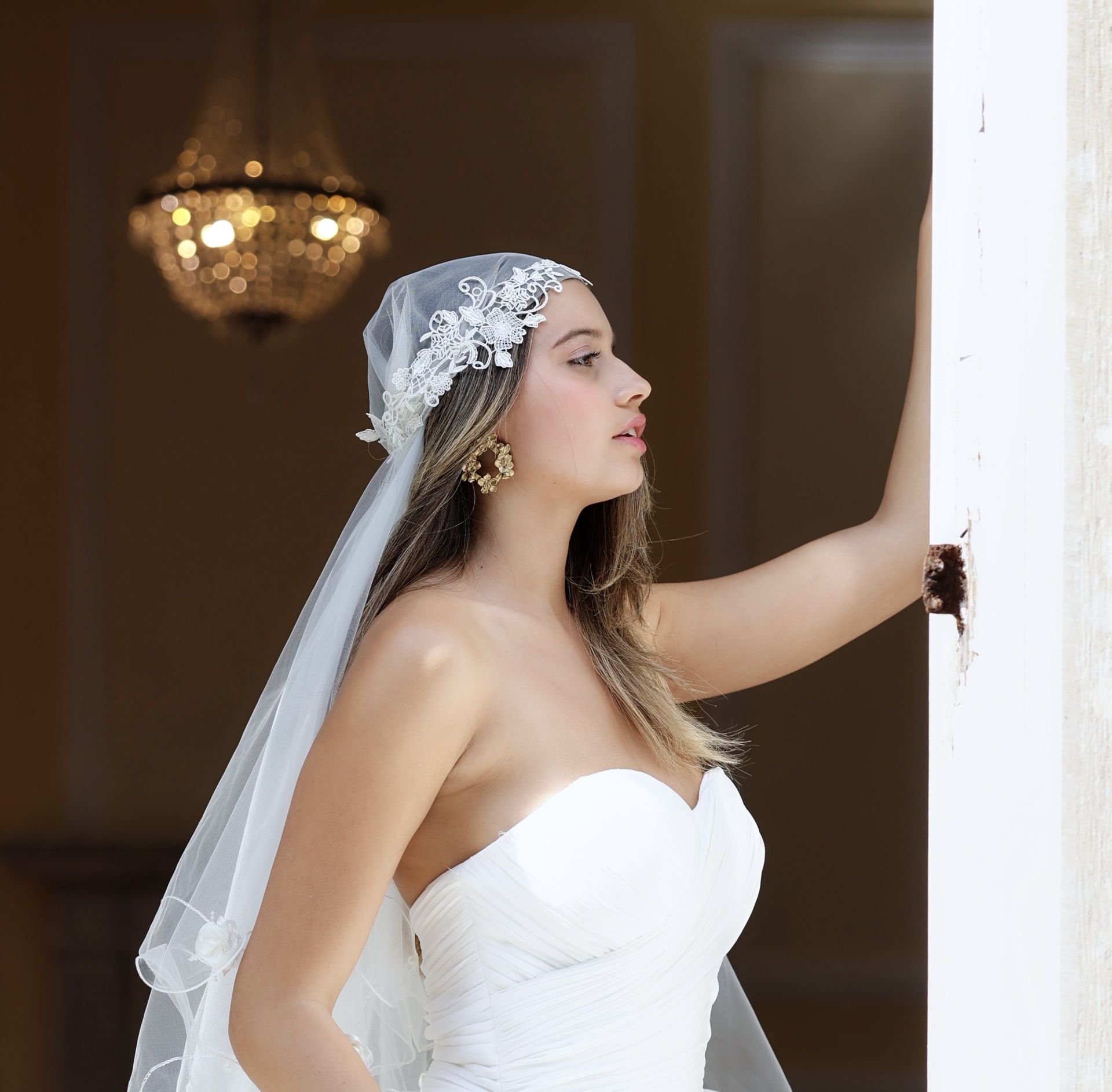 Trinity Mulher com vestido e véu de noiva branco, rosto de perfil e brincos dourados, candelabro desfocado ao fundo.