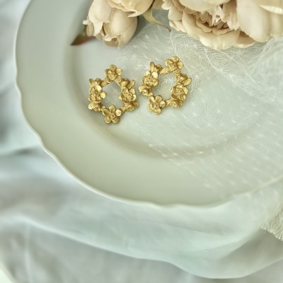Brincos dourados florais sobre prato branco com flores bege e tecido branco.