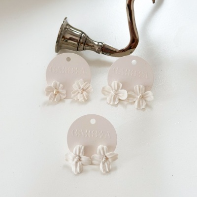 Três pares de brincos florais brancos em cartões CANOPA sobre superfície branca com suporte metálico