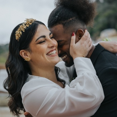 Casal abraçado, mulher com vestido branco e tiara dourada, homem com fato preto, fundo desfocado.