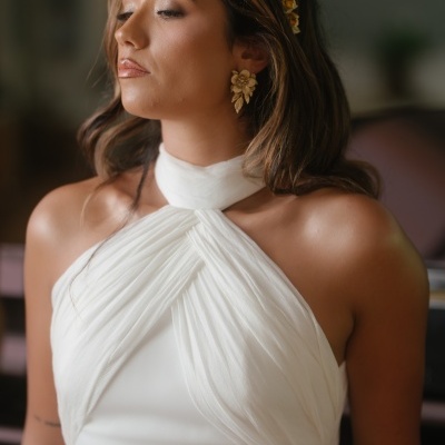 Mulher com vestido branco, brincos dourados e faixa dourada no cabelo num fundo desfocado