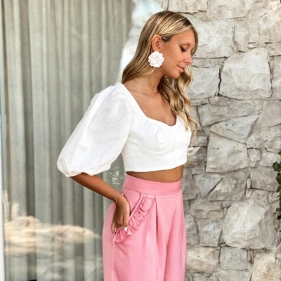 Mulher com blusa branca cropped de mangas bufantes e calças cor de rosa junto a parede de pedra