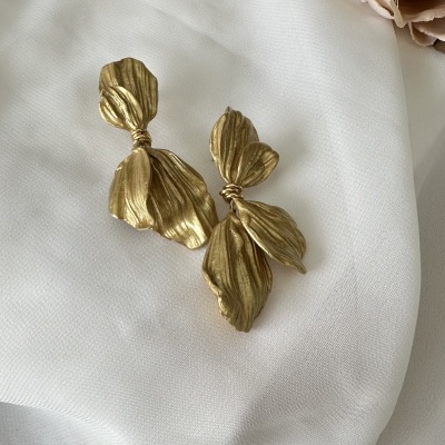 Brincos dourados de folhas sobre tecido branco com flores e folhas ao fundo