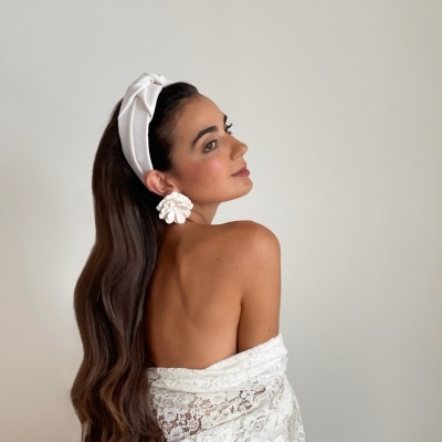 Mulher com turbante branco, brincos brancos grandes e xale de renda branco sobre fundo cinza claro