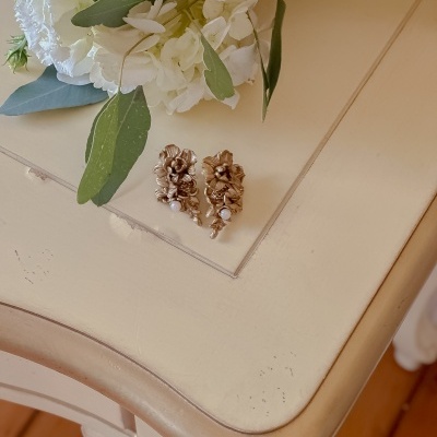 Brincos dourados em forma de flor com pérolas sobre mesa bege com flores brancas ao lado