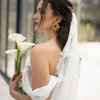 Noiva de perfil com vestido rendado branco, véu com renda, brincos dourados grandes e segurando flores brancas
