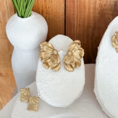 Brincos dourados em forma de flores em suporte branco com jarra branca e folhas verdes ao fundo