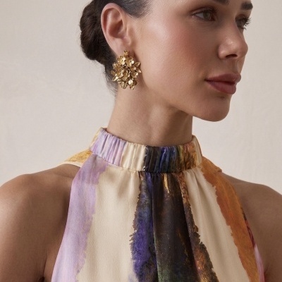 Brincos dourados florais grandes usados por mulher com vestido colorido e gola alta.