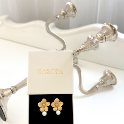 Brincos florais dourados com pérolas numa caixa GAROTA junto a castiçal prateado