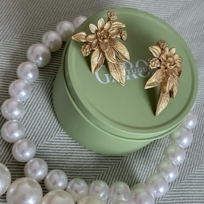 Brincos dourados florais com colar de pérolas sobre estojo verde e fundo em tecido cinza