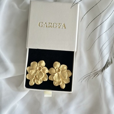 Brincos dourados em forma de flor numa caixa branca com interior preto e texto GAROTA